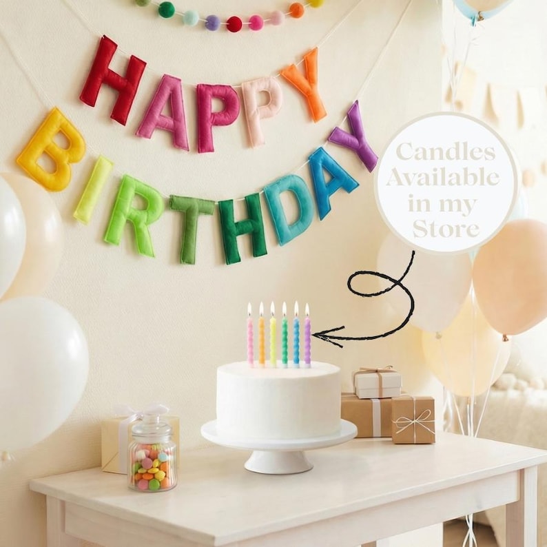 May include: A white birthday cake with lit rainbow candles on a white stand. A colourful "Happy Birthday" banner hangs above, with balloons and gifts on a white table. A jar of colourful sweets is also on the table.
