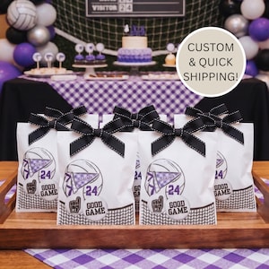 May include: White party favor bags with black bows, featuring a volleyball graphic with the text "Wildcats 24 Good Game". The bags are arranged on a wooden tray, with a purple and white checkered tablecloth.
