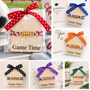 May include: A collection of bracelets packaged in clear plastic bags with polka dot ribbons. Each bracelet features gold-toned letters spelling words like OHIO, UK, EAST, VOLTS, KSU, or RHS, attached to woven bands in various colors matching the ribbons.