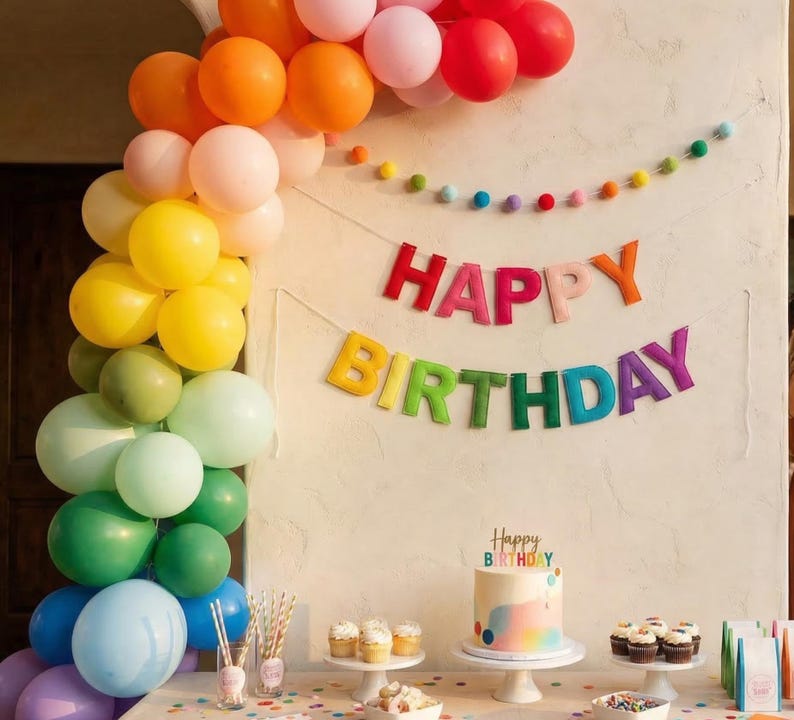 May include: A vibrant birthday celebration featuring a rainbow balloon arch, a 'Happy Birthday' banner, and a cake. The balloons are in shades of orange, yellow, green, blue, pink, and red. A cake with a rainbow design is on a white cake stand.