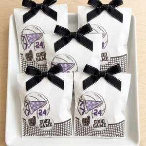 May include: White gift bags with black bows and stitching, arranged on a white tray. Each bag features a basketball graphic with the text "Wildcats", "24", and "Good Game". The bags are decorated with a black and white net design.