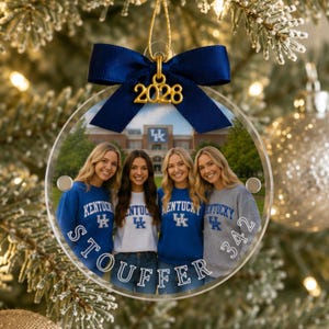 May include: A circular clear acrylic Christmas ornament featuring a photograph of four people wearing University of Kentucky apparel in front of a campus building. The ornament is topped with a dark blue satin bow and a gold 2028 charm.