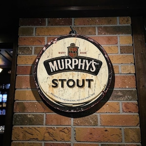 Puede incluir: Letrero metálico redondo y desgastado de Murphy's Stout, montado en una pared de ladrillo. El letrero presenta el logotipo de Murphy's en blanco y negro, con la palabra "STOUT" en negrita. También dice "BREWED TO THE ORIGINAL RECIPE SINCE 1856."