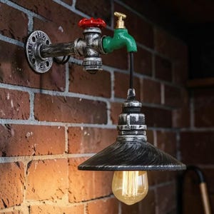 Può includere: Un'esclusiva lampada da parete progettata per assomigliare a un impianto idraulico. L'apparecchio presenta un paralume nero e argento con una lampadina in stile vintage, collegato a una struttura a forma di rubinetto con accenti rossi e verdi. Montato su un muro di mattoni.