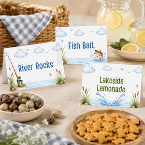 Könnte beinhalten: Drei weiße, gefaltete Tischkarten mit Aquarellillustrationen von Wolken, Wasser und Schilf. Die Karten zeigen den Text River Rocks, Fish Bait und Lakeside Lemonade in blauen und grünen Schriftarten mit grafischen Angelmotiven.