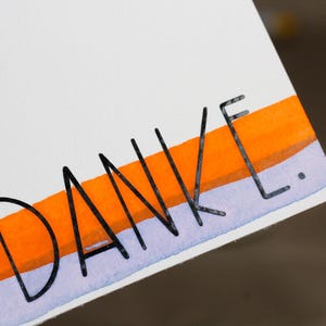 May include: A white card with the word "DANKE" in black lettering. The word is above a horizontal band of orange and light purple watercolor paint.