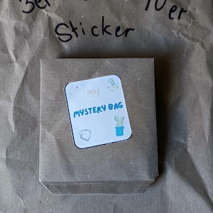 Könnte beinhalten: Ein brauner Papiersack mit einem Aufkleber mit der Aufschrift "MYSTERY BAG" in Blau. Der Aufkleber zeigt kleine Illustrationen eines Herzens, eines Bären, eines Dinosauriers, eines Kaktus und eines Hemdes. Der Text "3er 5er 10er Sticker" steht über der Tasche.