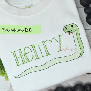 Könnte beinhalten: Weißes Langarmshirt mit einem grün gestickten Schlangenmuster. Der Name "Henry" ist grün gestickt, mit dem Text "Font not included" darüber. Die Schlange hat eine rote Zunge.
