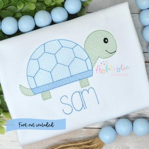 May include: White shirt featuring a light blue turtle design. The turtle has a blue shell and green head and feet. The name "Sam" is embroidered in blue. The text "Font not included" is in a light blue banner.