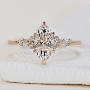 Puede incluir: Un anillo de compromiso de oro con un diamante central de talla marquesa. El anillo está adornado con diamantes redondos más pequeños y diamantes en forma de pera, engastados en una delicada banda de oro. El diseño del anillo es elegante.