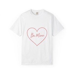 Peut inclure: T-shirt blanc avec un contour de cœur rouge et les mots "Be Mine" en cursive à l'intérieur. Le t-shirt a des manches courtes et un col rond.