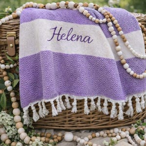 May include: A woven purple and white beach towel with the name "Helena" embroidered on it, draped over a wicker basket. The towel has a diamond pattern and white fringe. Wooden beads and decorative flowers surround the basket.
