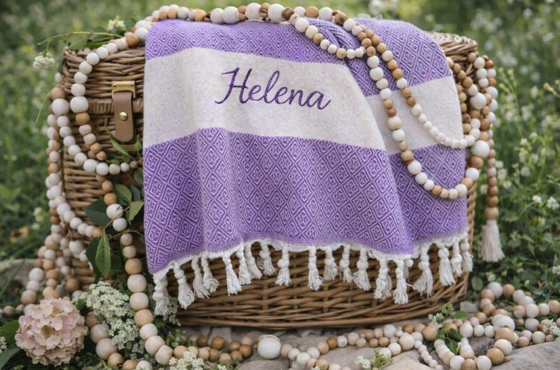 May include: A woven purple and white beach towel with the name "Helena" embroidered on it, draped over a wicker basket. The towel has a diamond pattern and white fringe. Wooden beads and decorative flowers surround the basket.