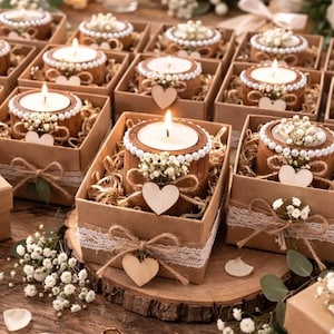 May include: A collection of lit candles in decorative wooden holders, each nestled in a small cardboard box. The candles are surrounded by white pearls and small white flowers, with a wooden heart and twine bow. The boxes are arranged on a wooden surface.