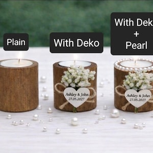 May include: Three wooden candle holders with lit candles. One is plain, one has a floral arrangement and a heart-shaped tag with text, and the third has pearls. The text on the tag reads "Ashley & John 27.05.2027".