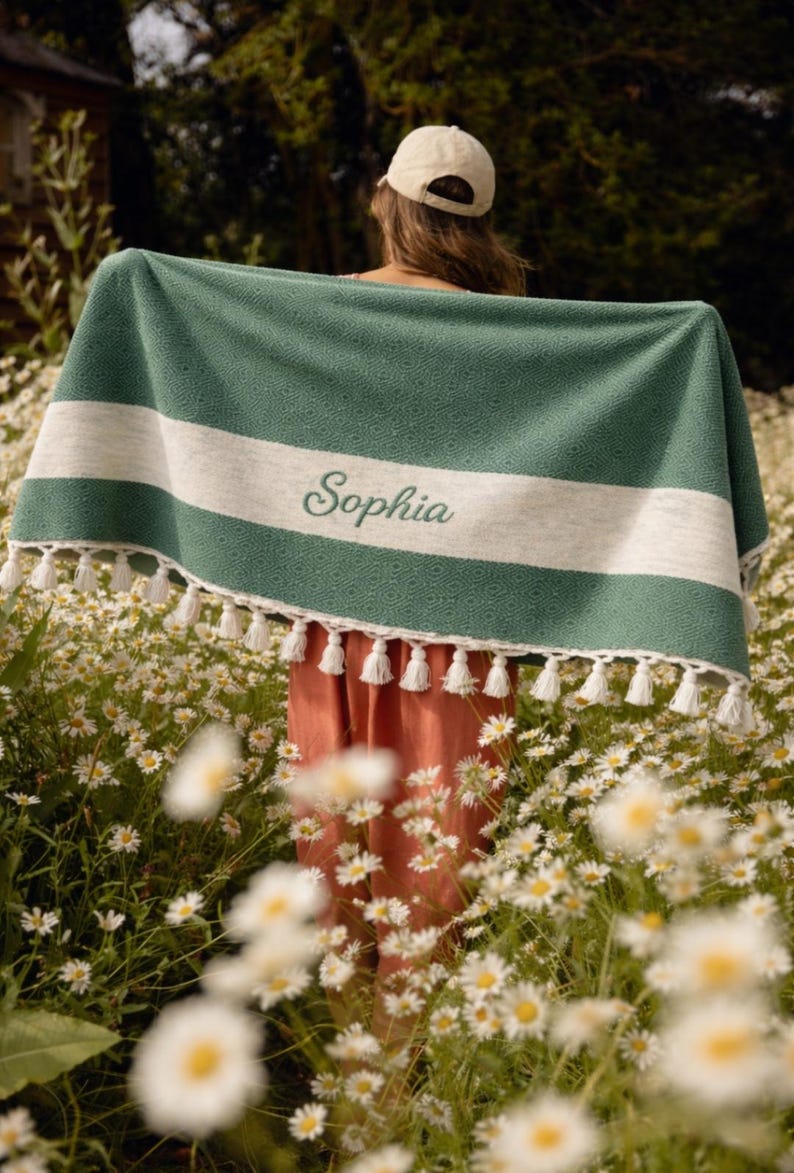 May include: A green and white personalized blanket with the name "Sophia" embroidered on it. The blanket has a diamond pattern and white tassels along the bottom edge. The person holding the blanket is standing in a field of daisies.