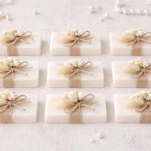 May include: Nine rectangular white soaps, each decorated with a small floral arrangement and a twine bow. The soaps are arranged in a grid, with scattered pearls and pearl strands for decoration.
