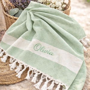 May include: A light green beach towel with white stripes and fringe. The towel has the name "Olivia" embroidered in green. A woven basket filled with flowers is in the background.