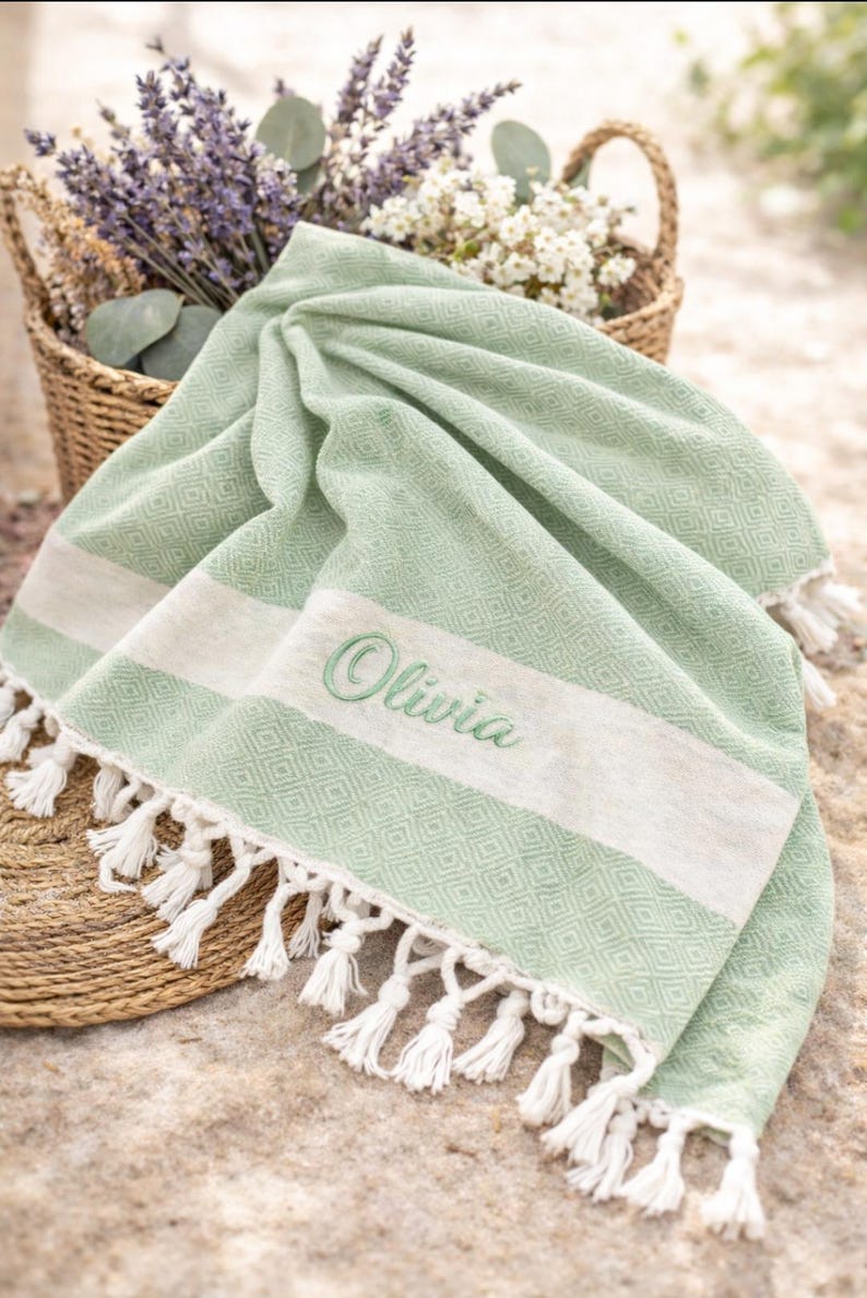 May include: A light green beach towel with white stripes and fringe. The towel has the name "Olivia" embroidered in green. A woven basket filled with flowers is in the background.