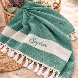 May include: A green and white beach towel with a diamond pattern and white tassels. The word "Sophia" is embroidered in cursive. A basket of flowers is in the background.