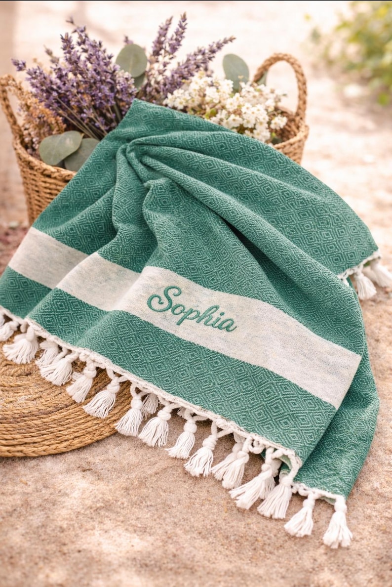 May include: A green and white beach towel with a diamond pattern and white tassels. The word "Sophia" is embroidered in cursive. A basket of flowers is in the background.
