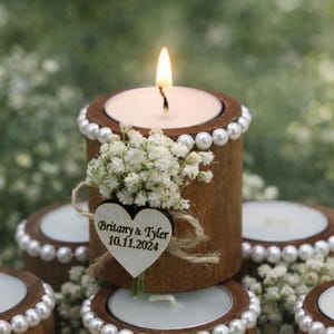 May include: A lit candle in a wooden tea light holder with a pearl trim and a small bouquet of white flowers. A wooden heart with the names "Britany & Tyler" and the date "10.11.2024" is attached. The candle is surrounded by other similar tea light holders.
