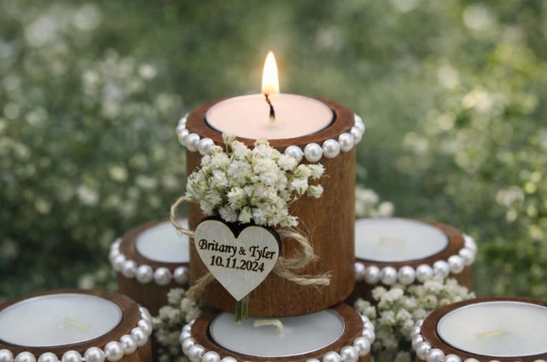 May include: A lit candle in a wooden tea light holder with a pearl trim and a small bouquet of white flowers. A wooden heart with the names "Britany & Tyler" and the date "10.11.2024" is attached. The candle is surrounded by other similar tea light holders.