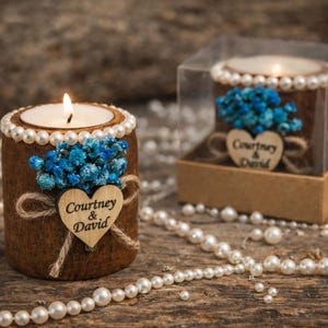 May include: A lit candle in a brown cylindrical holder decorated with pearls, blue flowers, and a wooden heart with the names "Courtney & David." The candle is on a wooden surface with scattered pearls.