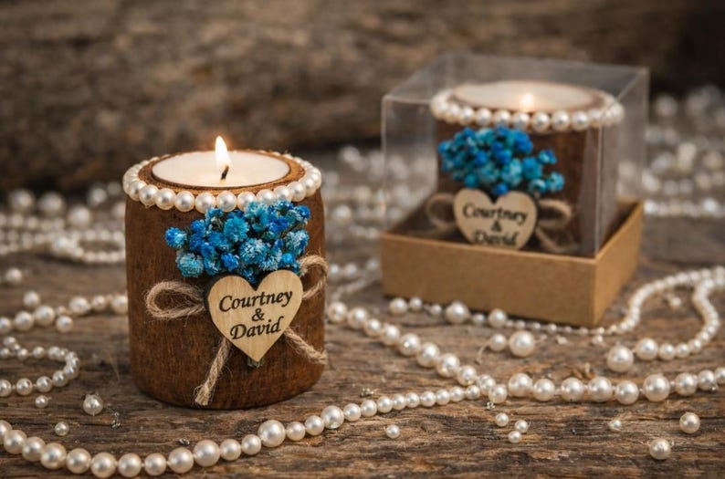 May include: A lit candle in a brown cylindrical holder decorated with pearls, blue flowers, and a wooden heart with the names "Courtney & David." The candle is on a wooden surface with scattered pearls.