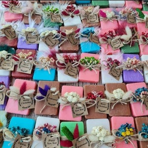 May include: A selection of colourful, rectangular soaps, each decorated with dried flowers, feathers, and twine. Small tags with text such as "Happy New Year", "Merry Christmas", and "Bridesmaid" are attached to some of the soaps.