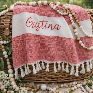 May include: A red and white patterned beach towel with the name "Cristina" embroidered on it, draped over a wicker basket. The towel has a fringe along the bottom. Wooden beads and flowers surround the basket.
