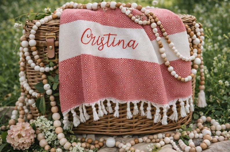 May include: A red and white patterned beach towel with the name "Cristina" embroidered on it, draped over a wicker basket. The towel has a fringe along the bottom. Wooden beads and flowers surround the basket.