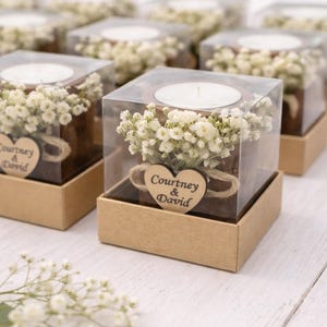 May include: Square candle holders with white candles, small white flowers, and a wooden heart with the text "Courtney & David". The candle holders are in clear boxes and set in brown boxes.