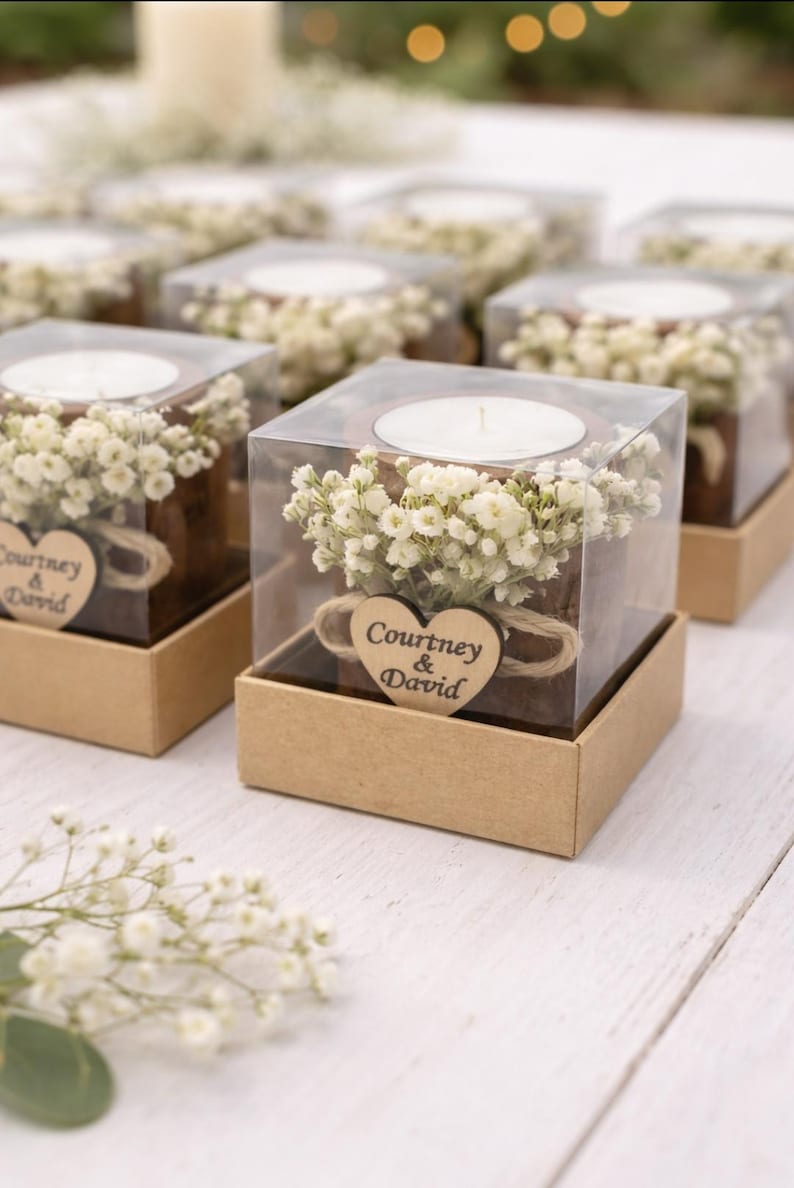 May include: Square candle holders with white candles, small white flowers, and a wooden heart with the text "Courtney & David". The candle holders are in clear boxes and set in brown boxes.