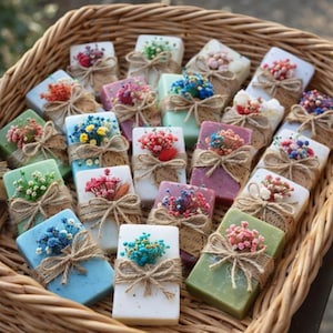 May include: A wicker basket filled with rectangular soaps in various colors, each adorned with dried flowers and tied with twine. The soaps are white, green, blue, pink, and purple, creating a colorful display.