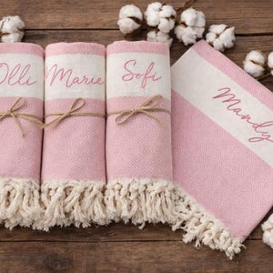 May include: Pink and white personalized towels with fringe. Three rolled towels are tied with twine and have names embroidered on them: Olli, Marie, and Sofi. A fourth towel is folded and reads Mandy.
