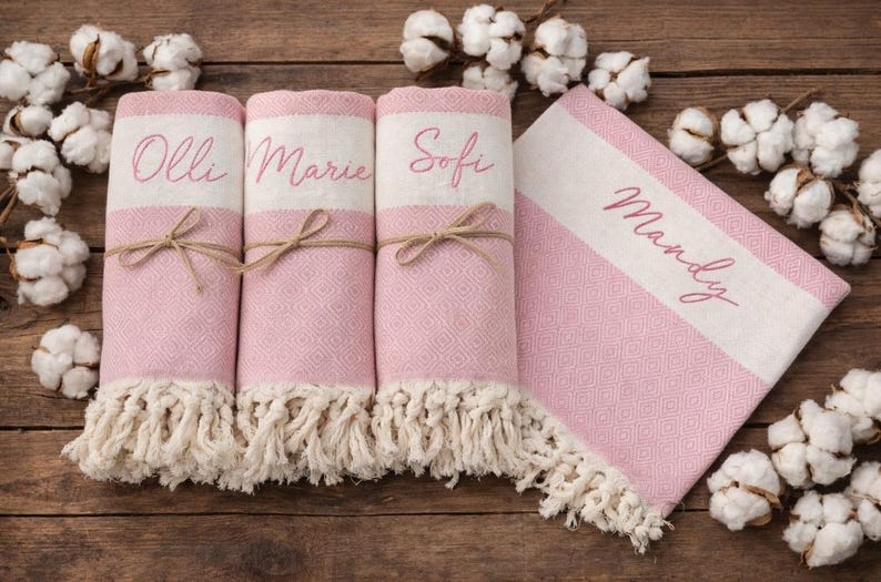 May include: Pink and white personalized towels with fringe. Three rolled towels are tied with twine and have names embroidered on them: Olli, Marie, and Sofi. A fourth towel is folded and reads Mandy.