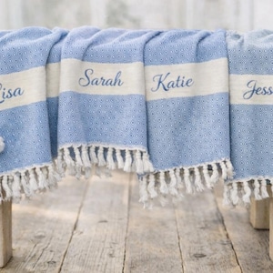 May include: A blue and white patterned throw blanket with fringe, personalized with the names "Lisa", "Sarah", "Katie", and "Jessica". The blanket is draped over a wooden bench, with cotton stems on either side.