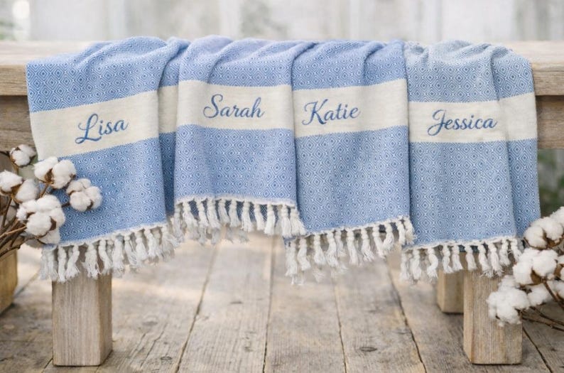May include: A blue and white patterned throw blanket with fringe, personalized with the names "Lisa", "Sarah", "Katie", and "Jessica". The blanket is draped over a wooden bench, with cotton stems on either side.