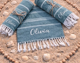 May include: A teal beach towel with white stripes and fringe, personalized with the name "Olivia" in white script. Two rolled towels are tied with twine. The scene is set on sand, surrounded by seashells and wooden beads.