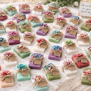May include: A collection of colourful soaps, each decorated with a small bouquet of dried flowers and a tag tied with twine. The soaps are in shades of white, green, purple, and red, arranged on a white surface.
