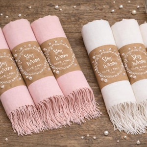 Könnte beinhalten: Gerollte rosa und weiße Schals mit Fransen, jeweils mit einem braunen Papierband umwickelt. Die Bänder sind mit Perlen und dem Text "Stay Warm" verziert. Verstreute Perlen und eine brennende Kerze sind im Hintergrund.