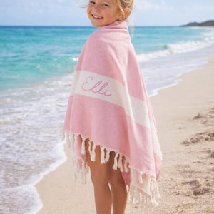 May include: A pink and white striped beach towel with the name "Elli" embroidered on it. The towel has a fringe trim and is wrapped around a person standing on a sandy beach. The background features the ocean and a blue sky.
