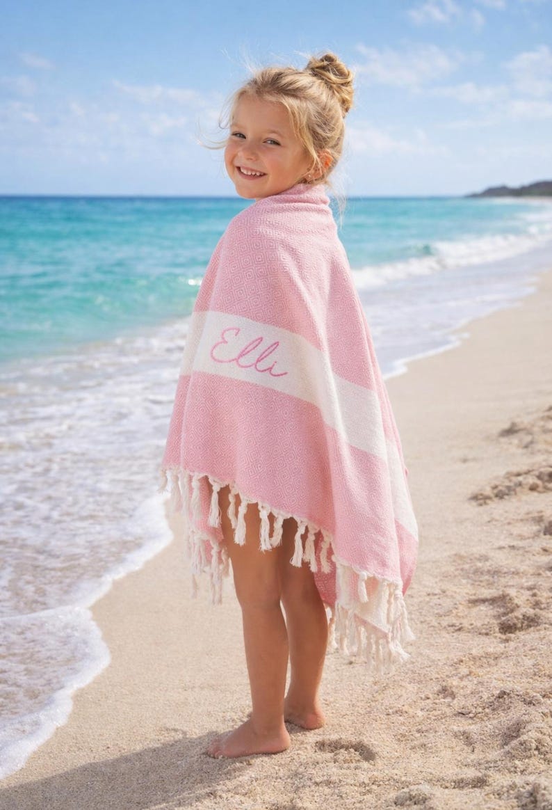 May include: A pink and white striped beach towel with the name "Elli" embroidered on it. The towel has a fringe trim and is wrapped around a person standing on a sandy beach. The background features the ocean and a blue sky.