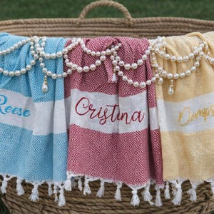 May include: Three personalized beach towels in a wicker basket, each with a name embroidered in a script font. The towels are blue, red, and yellow, with white stripes and diamond patterns. Pearl garlands adorn the basket's rim.