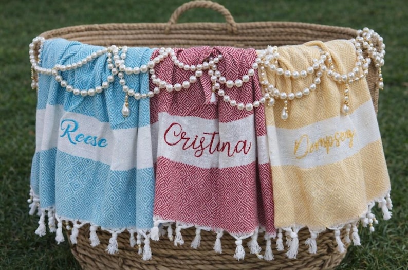 May include: Three personalized beach towels in a wicker basket, each with a name embroidered in a script font. The towels are blue, red, and yellow, with white stripes and diamond patterns. Pearl garlands adorn the basket's rim.