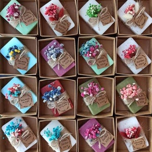 May include: A selection of decorative soaps in small cardboard boxes. Each soap is a different colour, wrapped with twine, and decorated with dried flowers and a small tag. The tags have handwritten text. The soaps are arranged in a grid pattern.