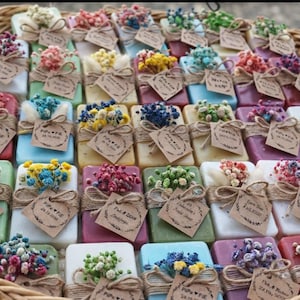 May include: A selection of colourful, rectangular soaps, each decorated with dried flowers and a small tag, presented in a woven basket. The soaps are in various colours, including pink, blue, green, and white, and are tied with twine.