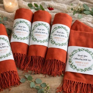 Puede incluir: Bufandas naranjas enrolladas con flecos, cada una con una etiqueta que dice "Thank You for Celebrating With Us! Hannah & Scott 6-16-2023." Se exhiben sobre una superficie de madera con elementos decorativos y una vela encendida.