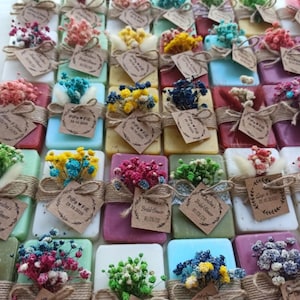 May include: A collection of colourful soaps, each decorated with dried flowers and tied with twine. Each soap has a small tag with text. The soaps are in various colours, including pink, blue, yellow, and green.